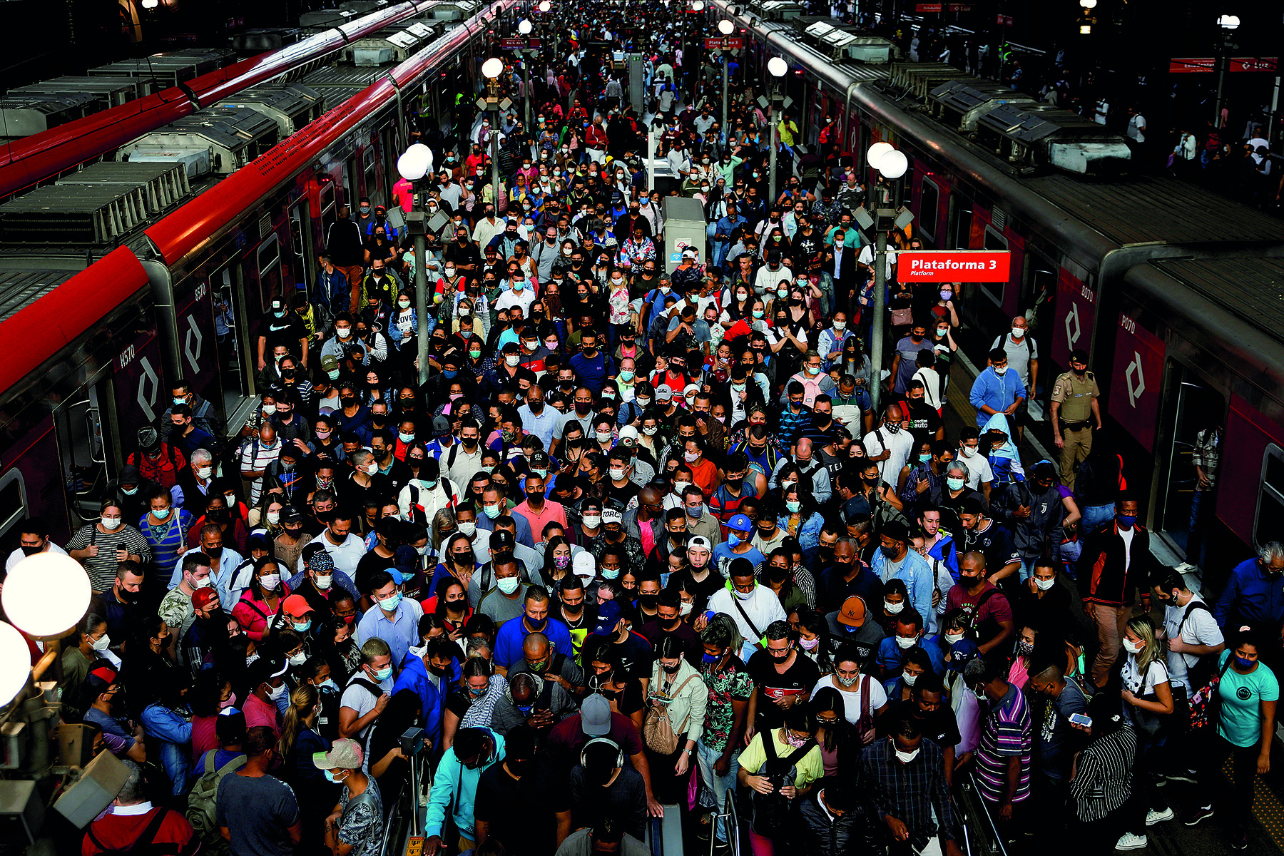 Fotografia. Uma multidão de pessoas espremidas em uma plataforma de estação de trem. Dois trens cinzas com linhas vermelhas estão parados com as portas abertas, um de cada lado da plataforma.