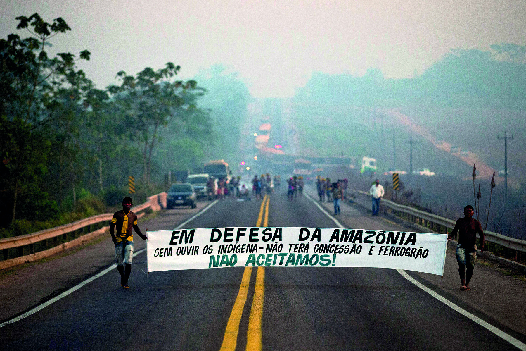 Fotografia. Imagem de dois homens indígenas em uma rodovia, segurando, um em cada ponta, uma faixa branca com os dizeres: 'Em defesa da Amazônia. Sem ouvir os indígenas não terá concessão e ferrogrão. Não aceitamos!'. Ao fundo, uma multidão está em pé junto à carros e caminhões, obstruindo a passagem.