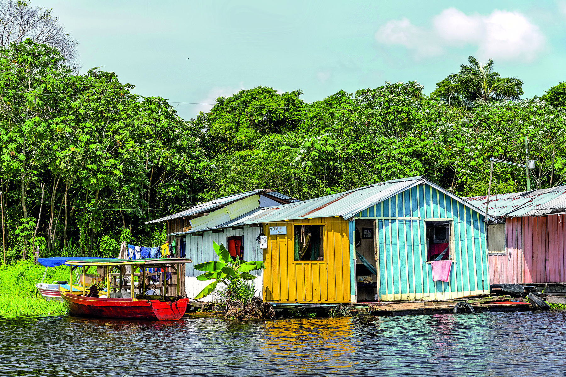 Fotografia. Imagem de casas de madeira coloridas construídas sobre as águas escuras de um rio. À esquerda, pequena embarcação. Ao fundo, vegetação verde e abundante.