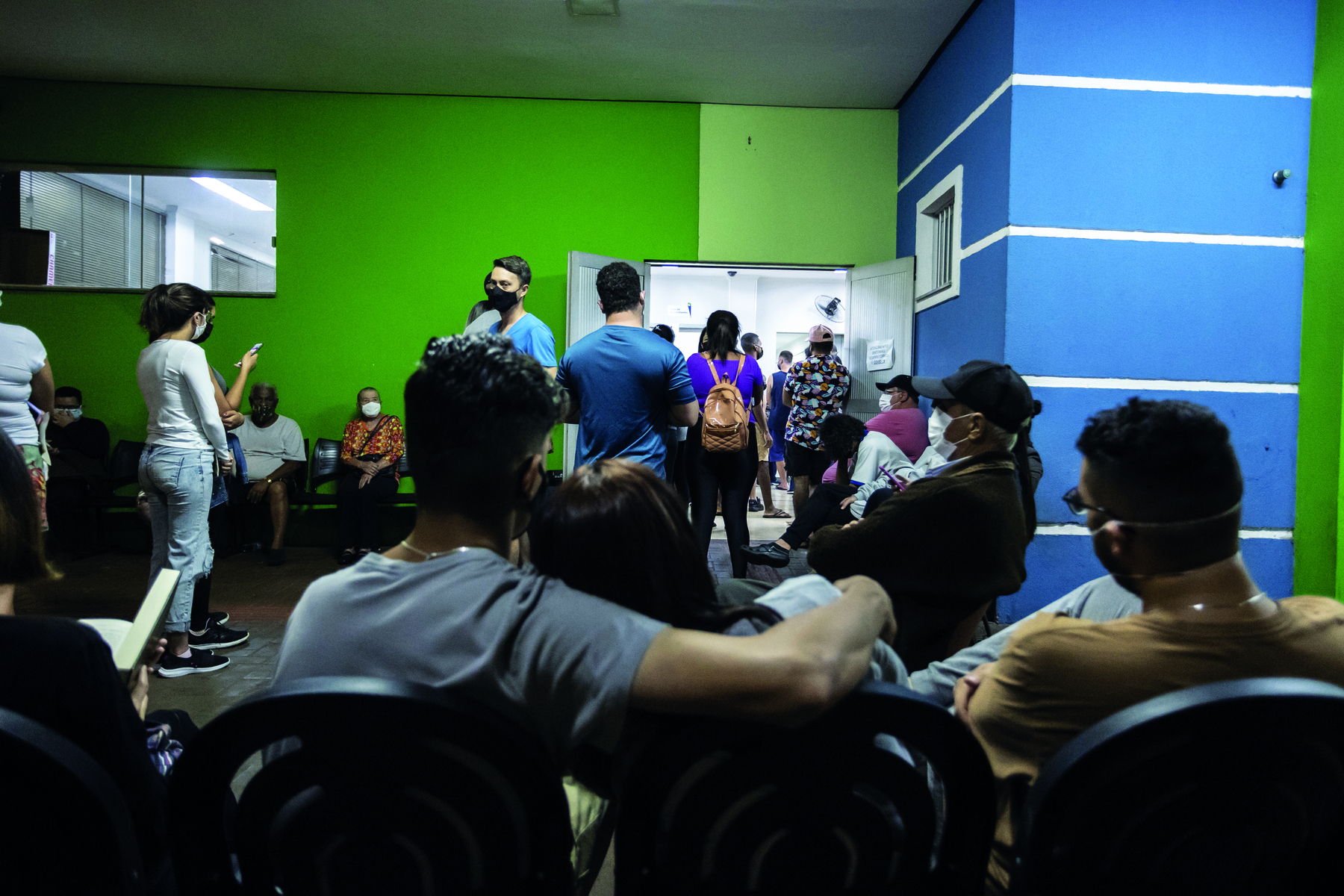 Fotografia. Em um espaço fechado, no primeiro plano, pessoas sentadas em cadeiras pretas e de costas para a câmera. À frente delas, há pessoas em pé em uma fila e, no fundo da sala, à esquerda, outras pessoas sentadas em cadeiras e, à direita, duas portas abertas dão acesso a outro ambiente.