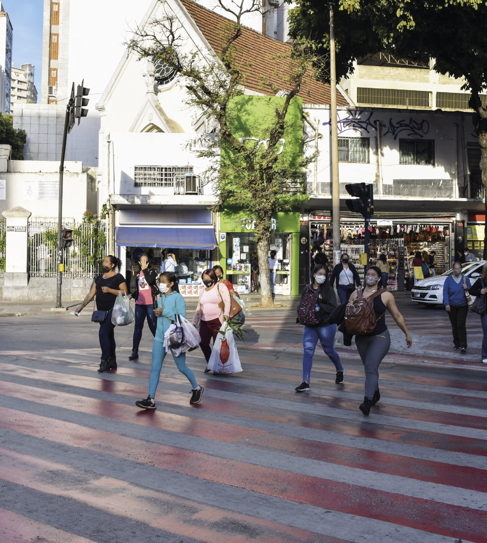 Fotografia. Em uma área aberta, pessoas usando máscaras de proteção para nariz e boca, carregando sacolas e mochilas, atravessam uma faixa de pedestres em primeiro plano. Ao fundo, árvore, carro, semáforo, poste e construções com dois andares, nas quais a parte térrea conta com estabelecimentos comerciais.