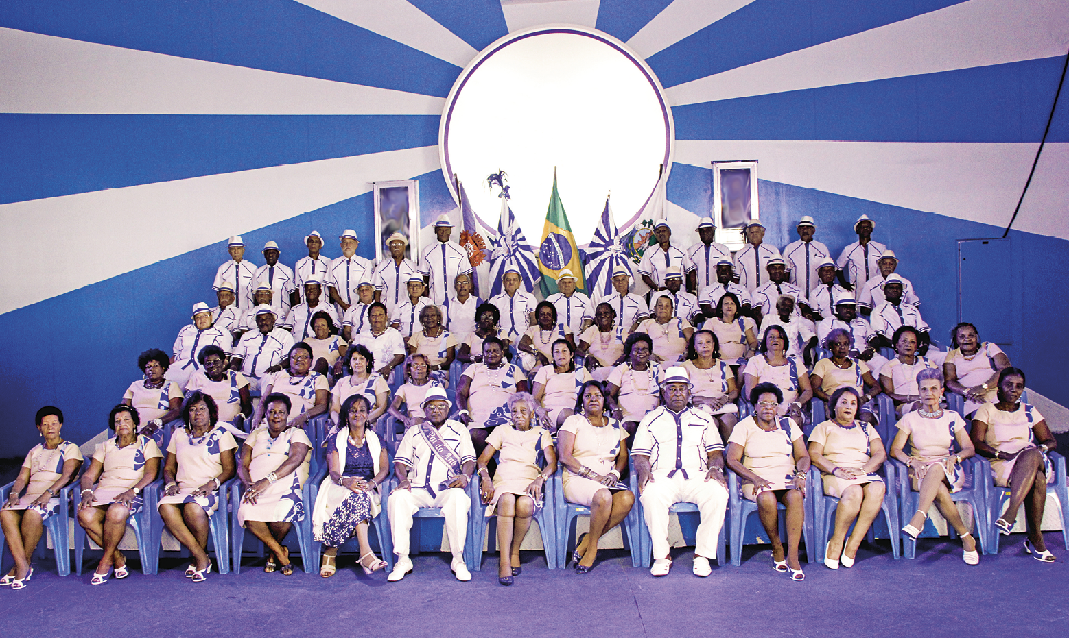 Fotografia. Pessoas uniformizadas e sentadas em cadeiras azuis. As mulheres usam um vestido bege com detalhes azuis e os homens camisas brancas com detalhes azuis e chapéu branco. No fundo, duas bandeiras em tons de branco e azul e, entre elas, a bandeira do Brasil. Também ao fundo, há um círculo e listras azuis e brancas radiais, que partem da borda do círculo.