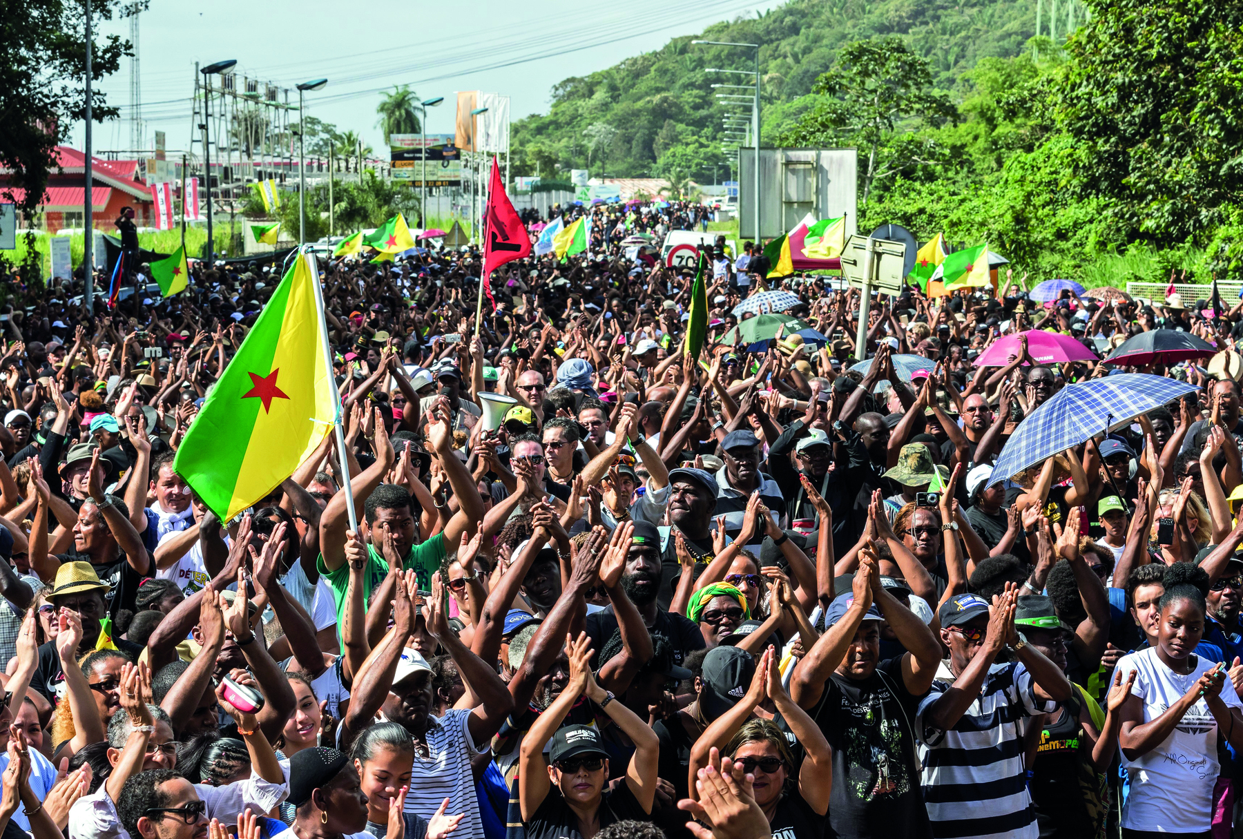 Fotografia. Uma multidão de pessoas em pé, com as mãos levantadas e juntas acima da cabeça. Há uma bandeira verde e amarela com uma estrela vermelha no centro; à direita, algumas pessoas seguram  guarda-chuvas abertos.