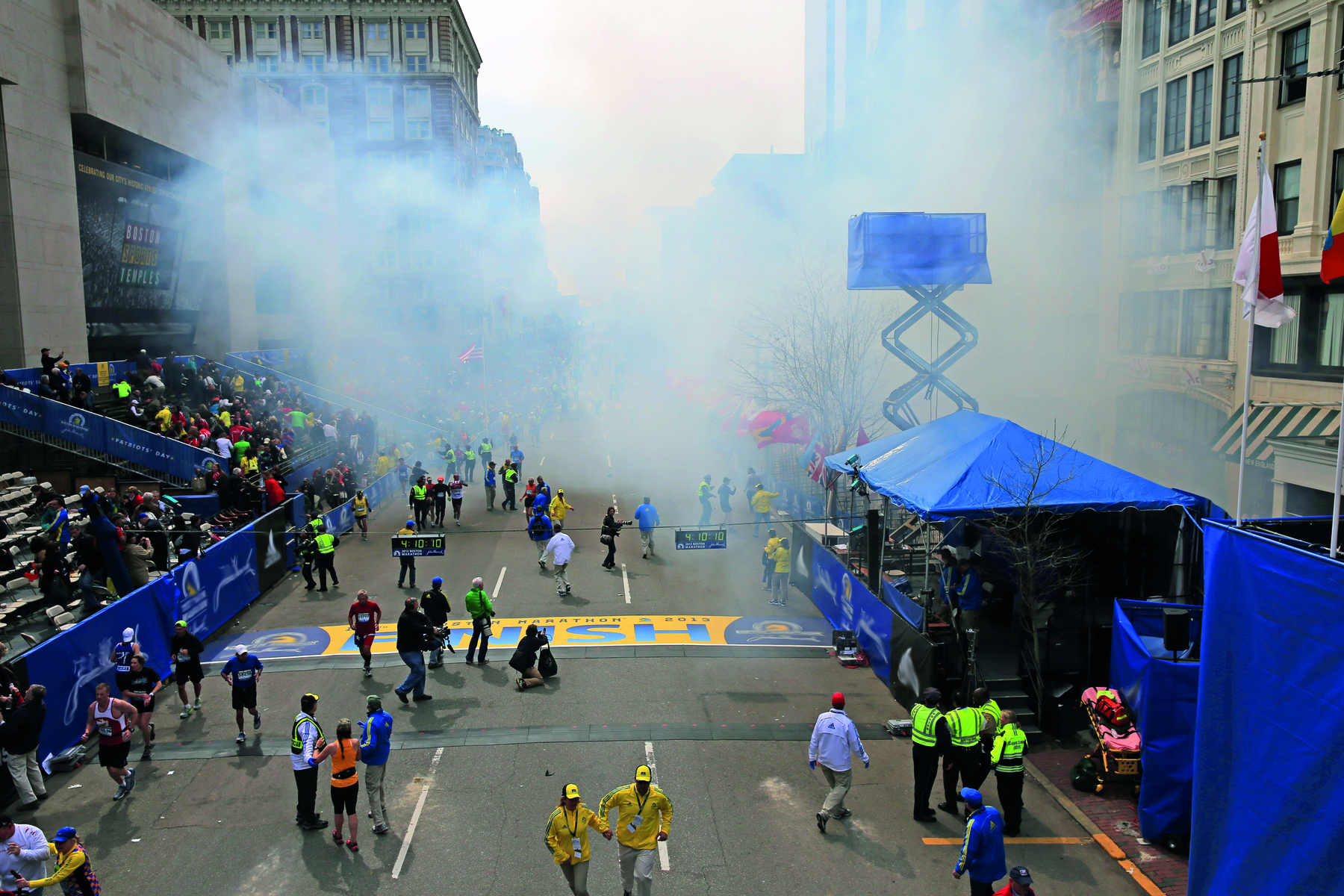 Fotografia. Vista aérea de uma rua com equipamentos azuis como barracas. Do lado esquerdo há uma arquibancada; no centro e nas laterais há diversas pessoas em pé. Ao fundo, uma cortina de fumaça branca.