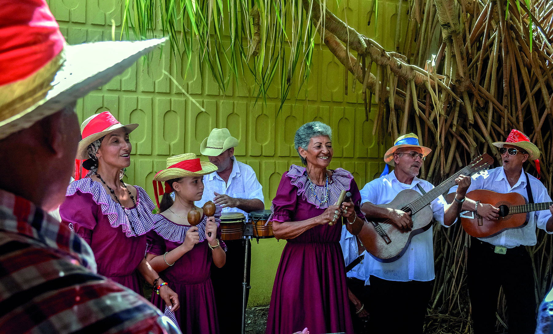 Fotografia. Pessoas tocando instrumentos musicais. Ao centro, uma mulher de cabelo grisalho e curto, usando vestido roxo, está segurando dois objetos de madeira e sorrindo; à esquerda, uma menina usando chapéu de palha, vestido roxo e segurando dois chocalhos; ao lado, outra mulher usando chapéu e vestido roxo; ambas estão sorrindo. À direita, dois homens usando chapéu de palha, camiseta branca, cada um segura um violão e estão com a boca aberta, cantando. Atrás da mulher que está no centro, um homem com chapéu de palha e camiseta branca toca dois pequenos tambores. Ao fundo, uma parede amarela e uma árvore.