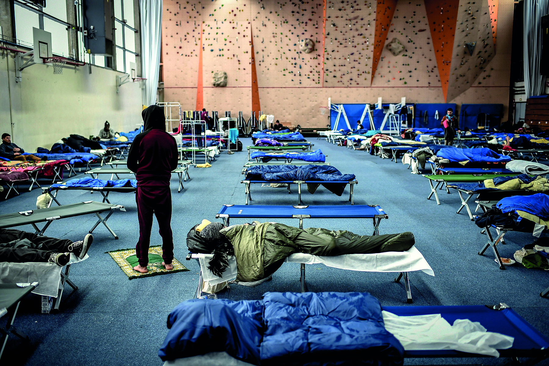 Fotografia. Interior de uma ginásio com diversas camas dobráveis e algumas pessoas deitadas e cobertas com cobertores cinzas ou azuis. No centro, uma pessoa em pé e de costas.