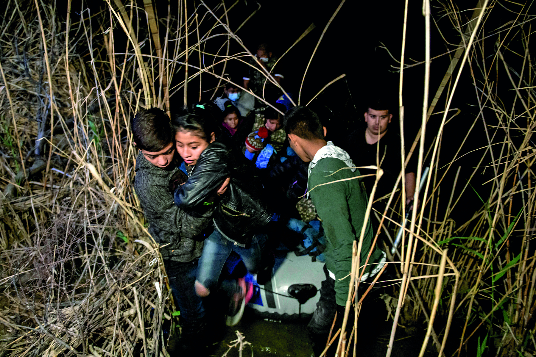 Fotografia. No meio de um matagal, a imagem retrata diversas pessoas passando por uma passagem entre a vegetação. O ambiente está escuro, com pouca iluminação, indicando que a situação ocorre à noite ou de madrugada. As pessoas maiores carregam e ajudam crianças e ao fundo outro grupo aguarda passagem.