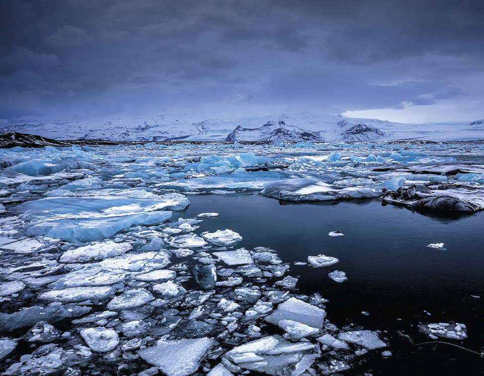 Fotografia. 
Imagem de uma geleira se decompondo em pequenos fragmentos brancos de gelo boiando sobre água de cor azul escuro. Ao fundo, uma superfície com colinas cobertas de neve e céu nublado.