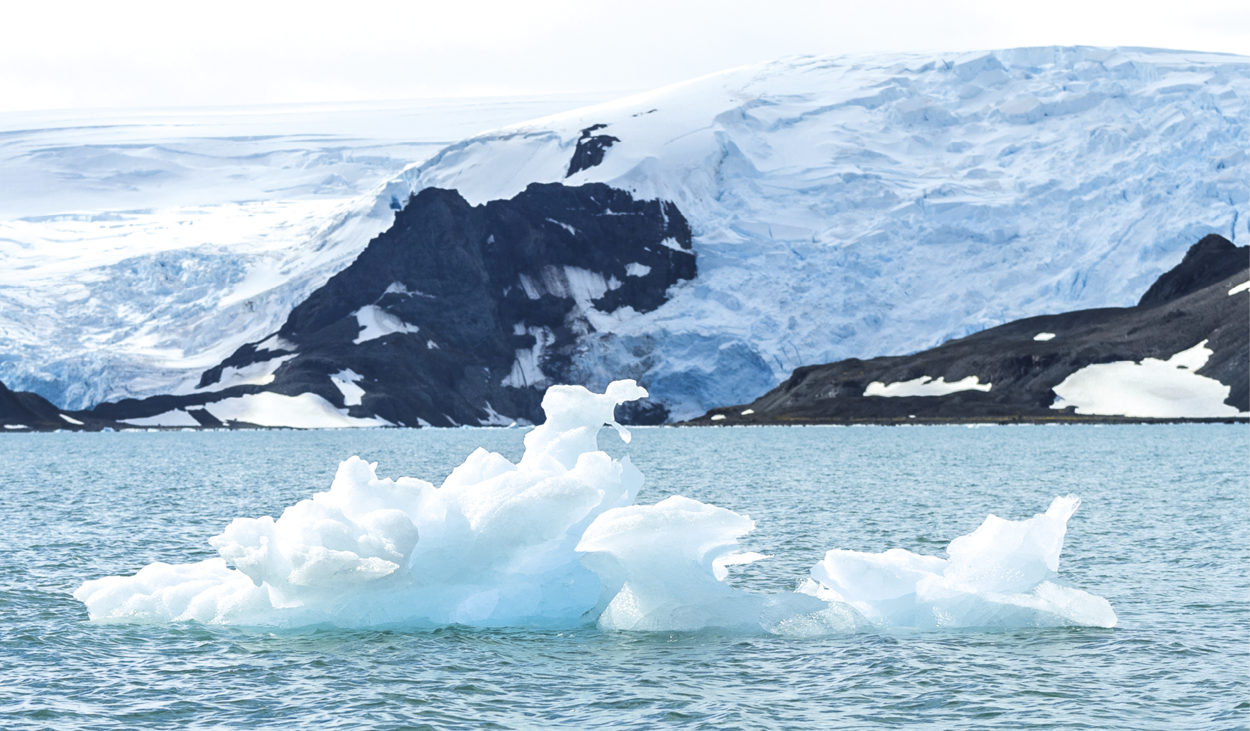 Fotografia. 
No primeiro plano, iceberg flutua na mar de águas azuis. Ao fundo, superfície montanhosa recoberta de gelo, exceto em algumas partes que têm solo exposto.