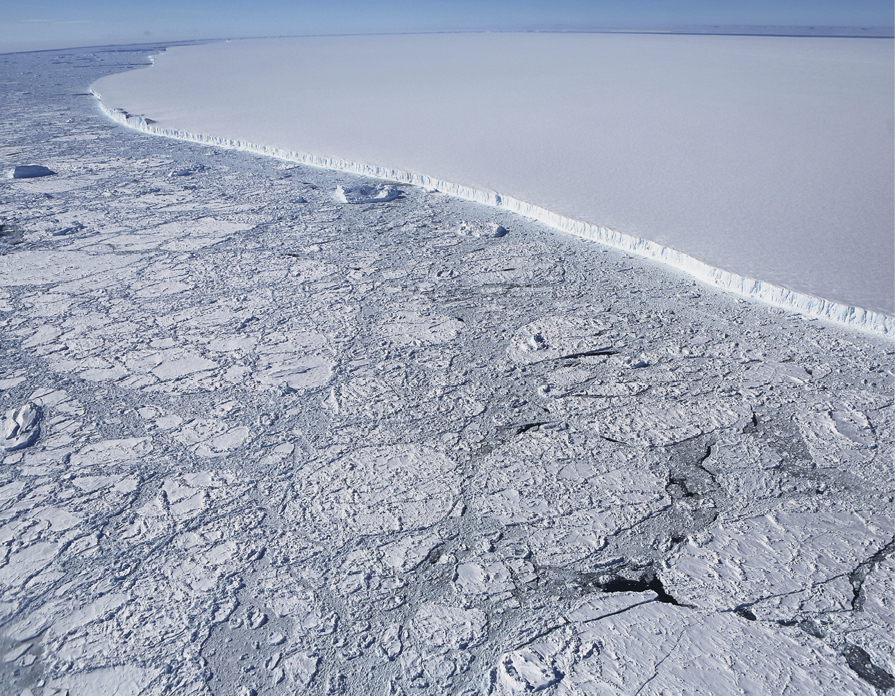 Fotografia. 
Vista aérea de extensa área de mar co placas de gelo flutuando e um grande iceberg de superfície plana e borda escarpada.