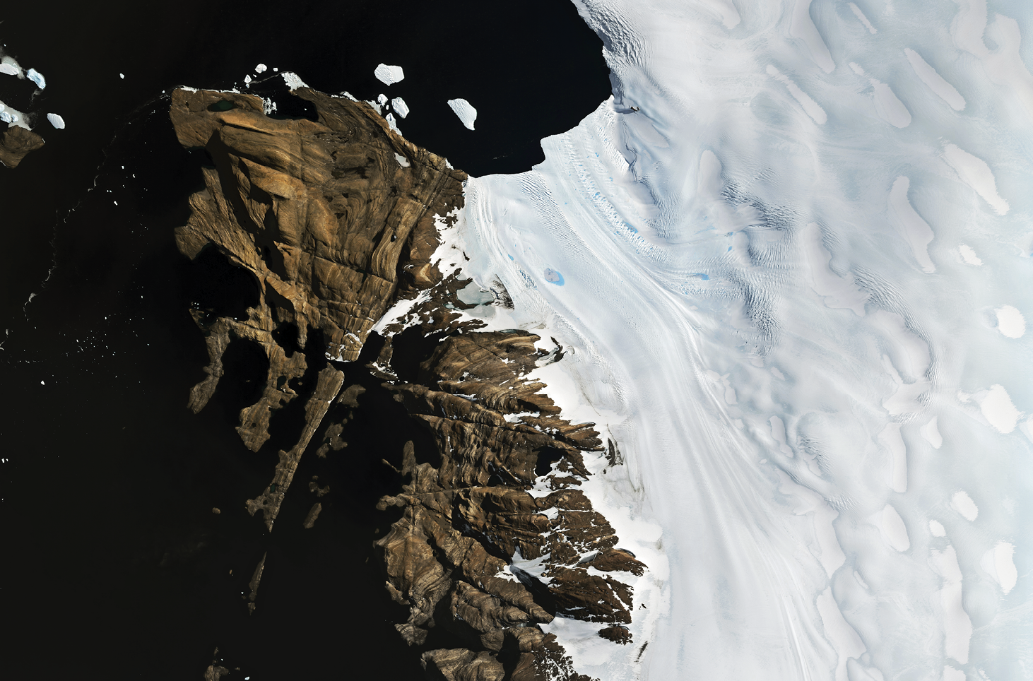 Imagem de satélite. 
Imagem de satélite destacando, à direita, uma grande área coberta de gelo, com uma linha de arrasto de gelo em direção ao mar. À esquerda, rochas expostas sem cobertura vegetal e glacial. Na parte superior esquerda, mar escuro com algumas placs de gelo.