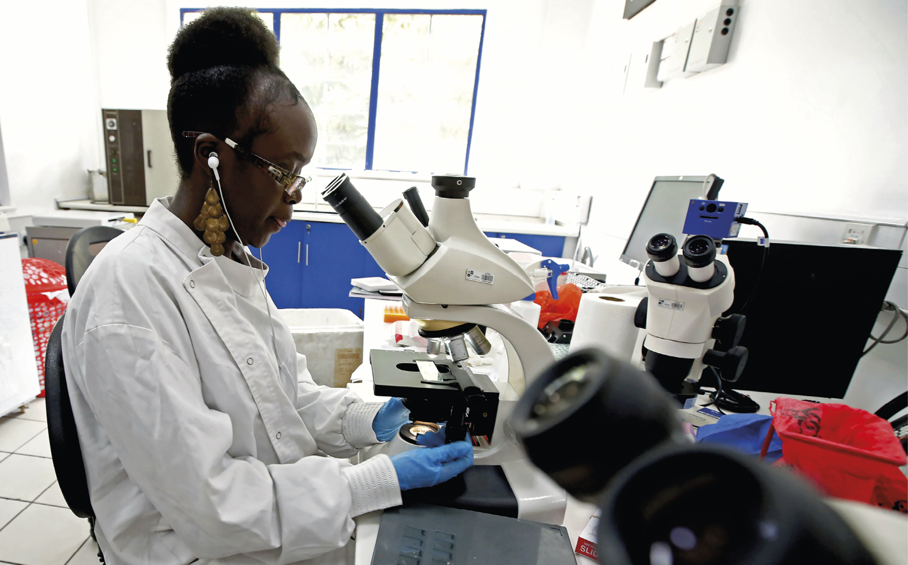 Fotografia. Destaque para uma mulher negra de avental branco e luvas azuis, fones de proteção auricular, cabelo preso e óculos de armação quadrada. Ela está sentada em um cadeira, dentro da sala de um laboratório, e utiliza um microscópio que está sobre uma bancada. Sobre a bancada há diversos equipamentos, entre eles um computador e outro microscópio. Ao fundo, armários baixos azuis e uma janela com iluminação natural que clareia o ambiente.