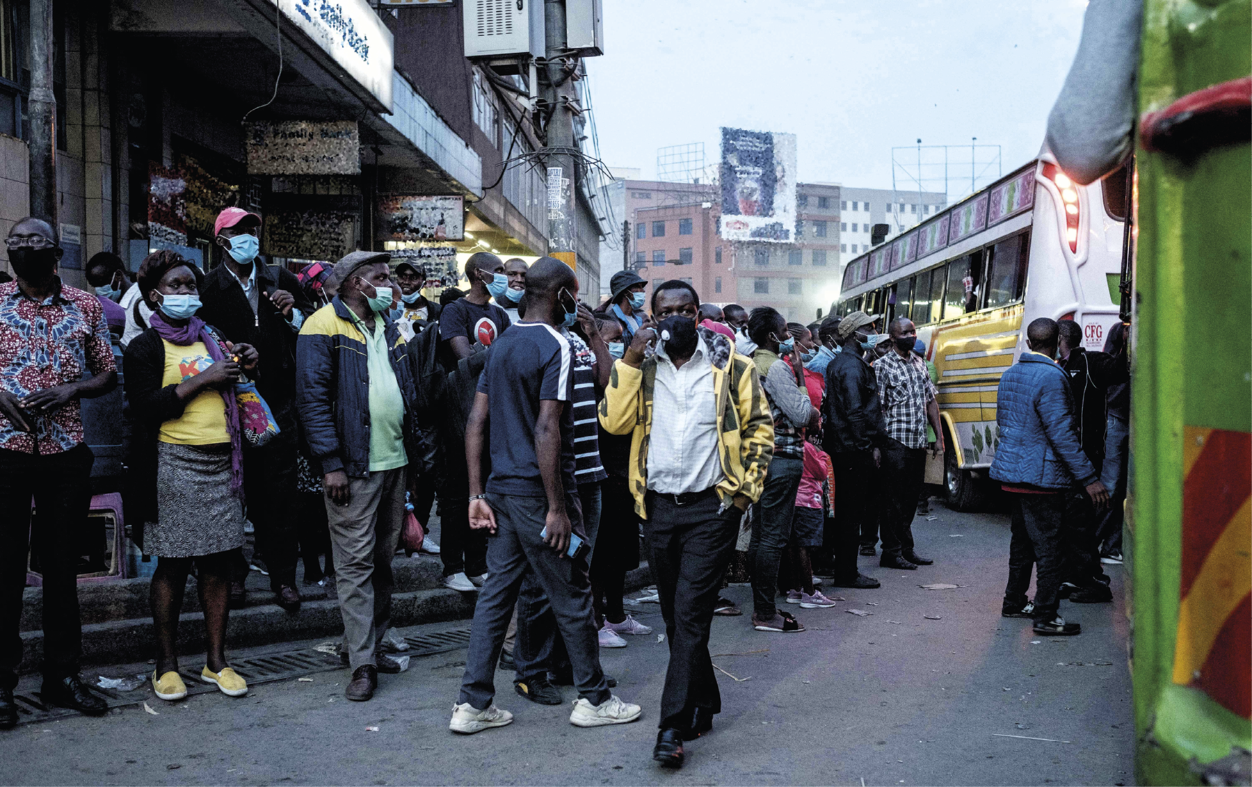 Fotografia. 
Pessoas com máscaras de proteção sobre a boca e o nariz estão aglomeradas em uma rua comercial. A maioria está parada na calçada; algumas andam pela rua. À direita, há um ônibus amarelo cercado de pessoas em um dos lados.