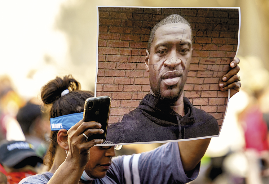 Fotografia. Uma mulher de cabeça baixa segura um celular em uma das mãos e, na outra, um cartaz grande com o rosto de um homem negro de cabelo curto e usando blusa preta.