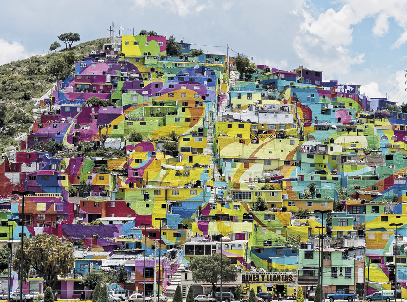 Fotografia. Vista, de baixo para cima, de um morro com diversas casas simples, baixas e em tons vibrantes de roxo, vermelho, lilás, azul, verde e amarelo, construídas muito próximas uma das outras até o topo. Do lado esquerdo da imagem, parte do morro aparece sem construções, com vegetação rasteira e algumas árvores.