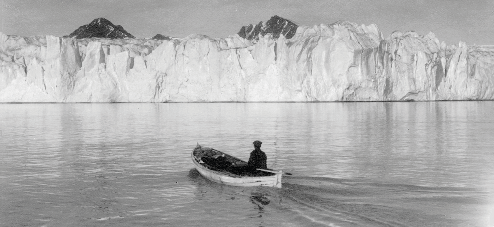 Fotografia. Em preto e branco. Uma pessoa está dentro de uma canoa em  águas calmas, navegando  em direção a uma geleira branca e alta, ao fundo. Atrás da geleira, topos de montes.