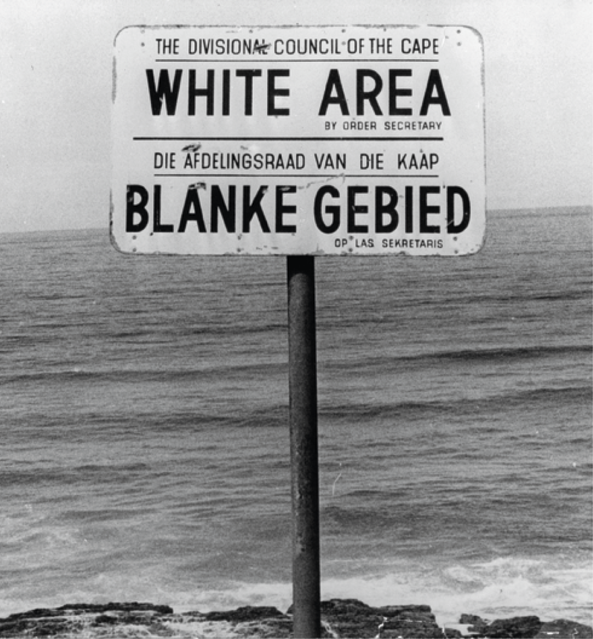 Fotografia. Em uma praia, fincada sobre uma uma rocha, às margens de águas marítimas, em destaque, uma placa na cor branca com um texto, na parte superior em inglês, na parte inferior em africâner, que significa: 'Área branca'.