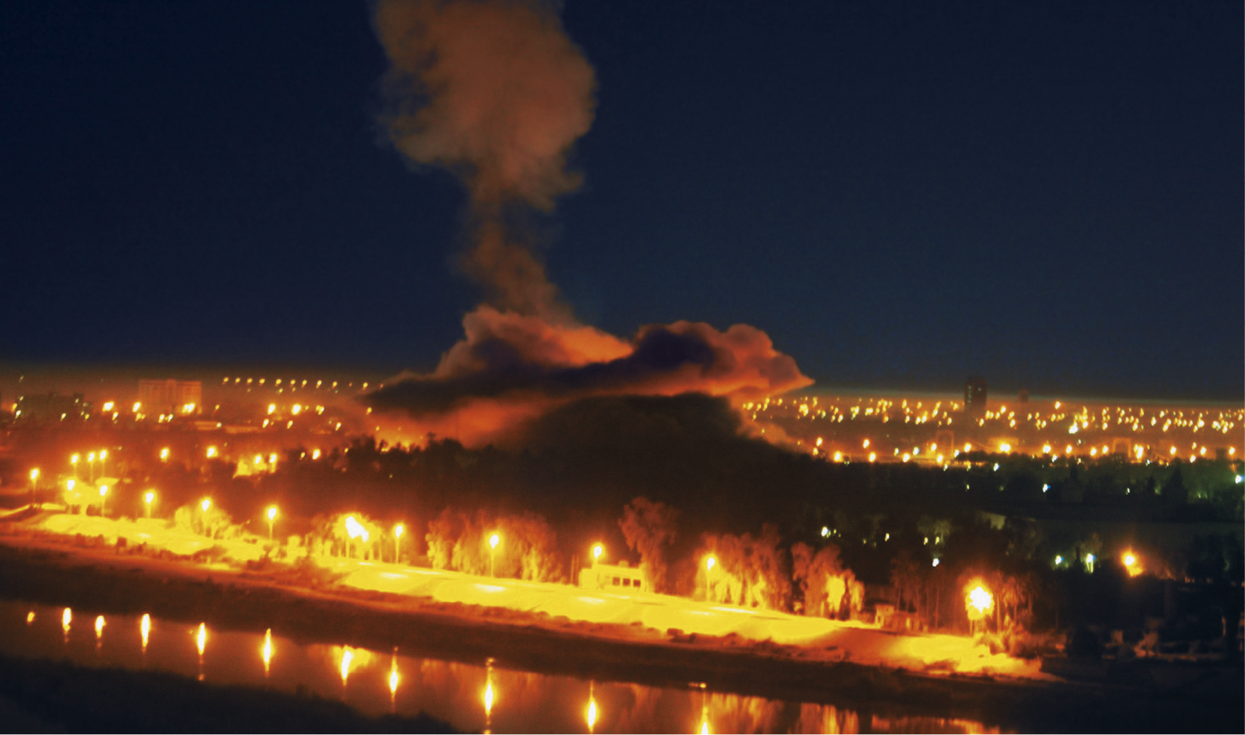 Fotografia. Vista aérea de uma coluna de fumaça alaranjada sobre uma cidade durante a noite. Ao redor há postes de luz e um rio.