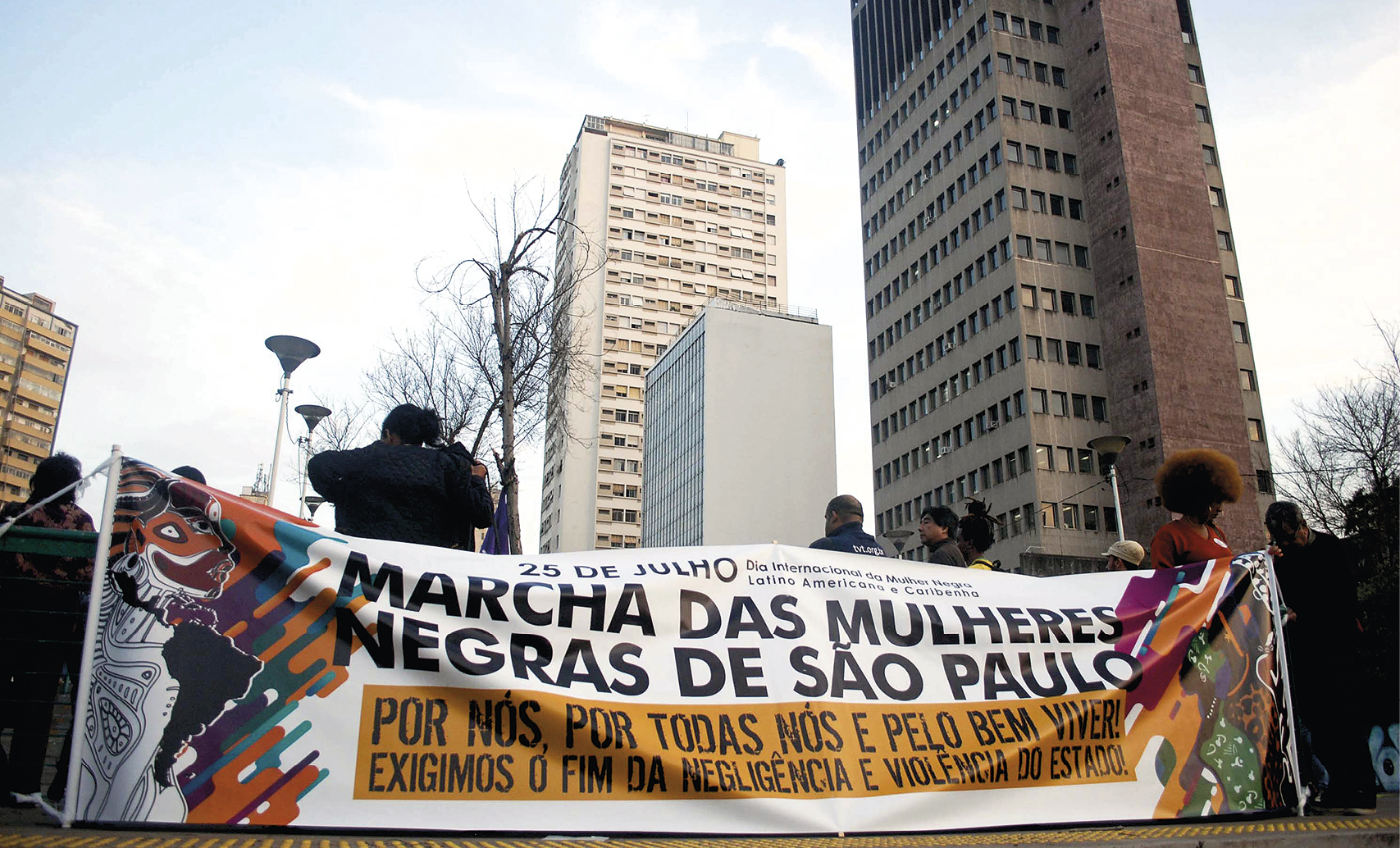 Fotografia. No primeiro plano, destaque para uma faixa de fundo branco com o texto: ‘’MARCHA DAS MULHERES NEGRAS DE SÃO PAULO – POR NÓS, POR TODAS NÓS E PELO BEM VIVER! EXIGIMOS O FIM DA NEGLIGÊNCIA E VIOLÊNCIA DO ESTADO’’. Atrás da faixa, algumas pessoas aparecem e, ao fundo, há prédios altos e postes de iluminação.