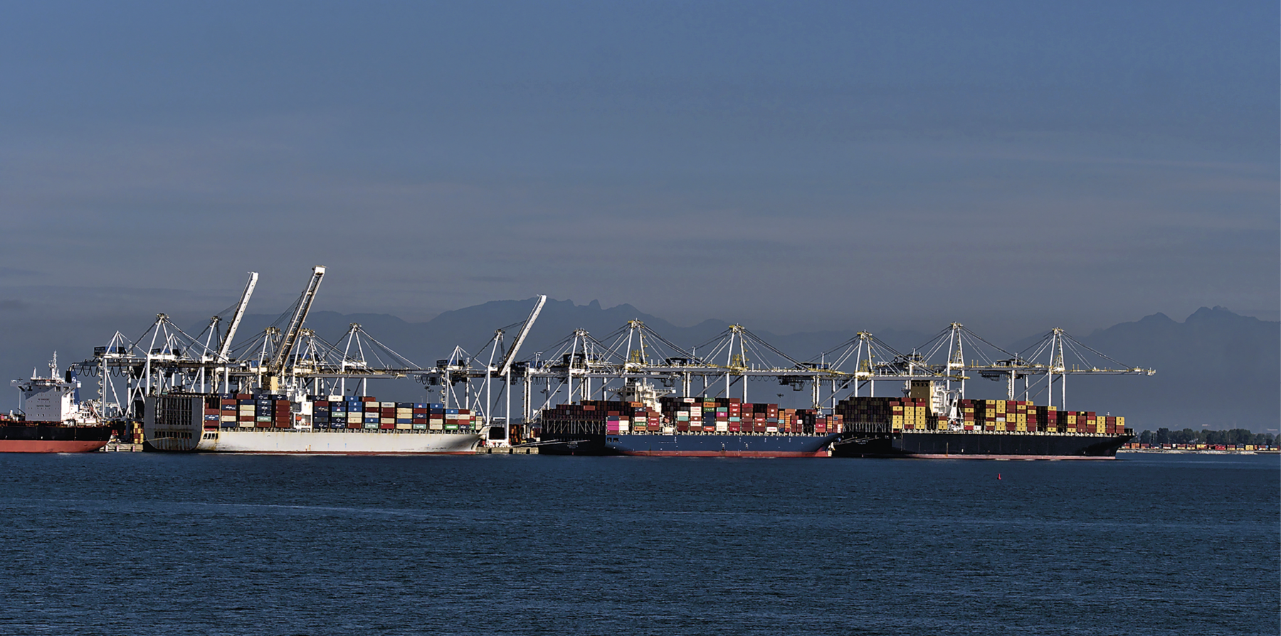 Fotografia. Vista de um porto oceânico com navios de carga ancorados. Os navios possuem grandes estruturas, como guindastes, e estão ocupados por diversos contêineres. Ao fundo montanhas e na parte superior da foto, céu azul.