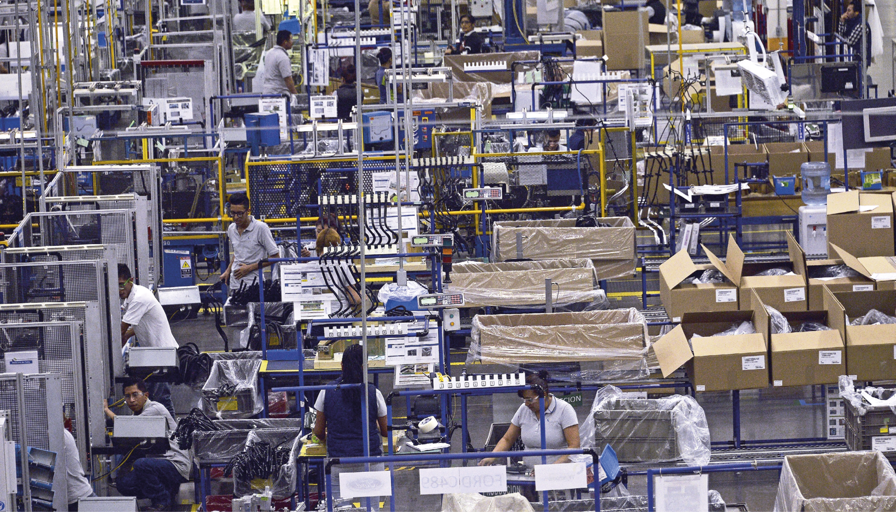 Fotografia. Parte interna de uma fábrica. Há vários corredores e posições de trabalho com pessoas uniformizadas operando máquinas. Do lado direito da posição das máquinas há caixas de papelão abertas.