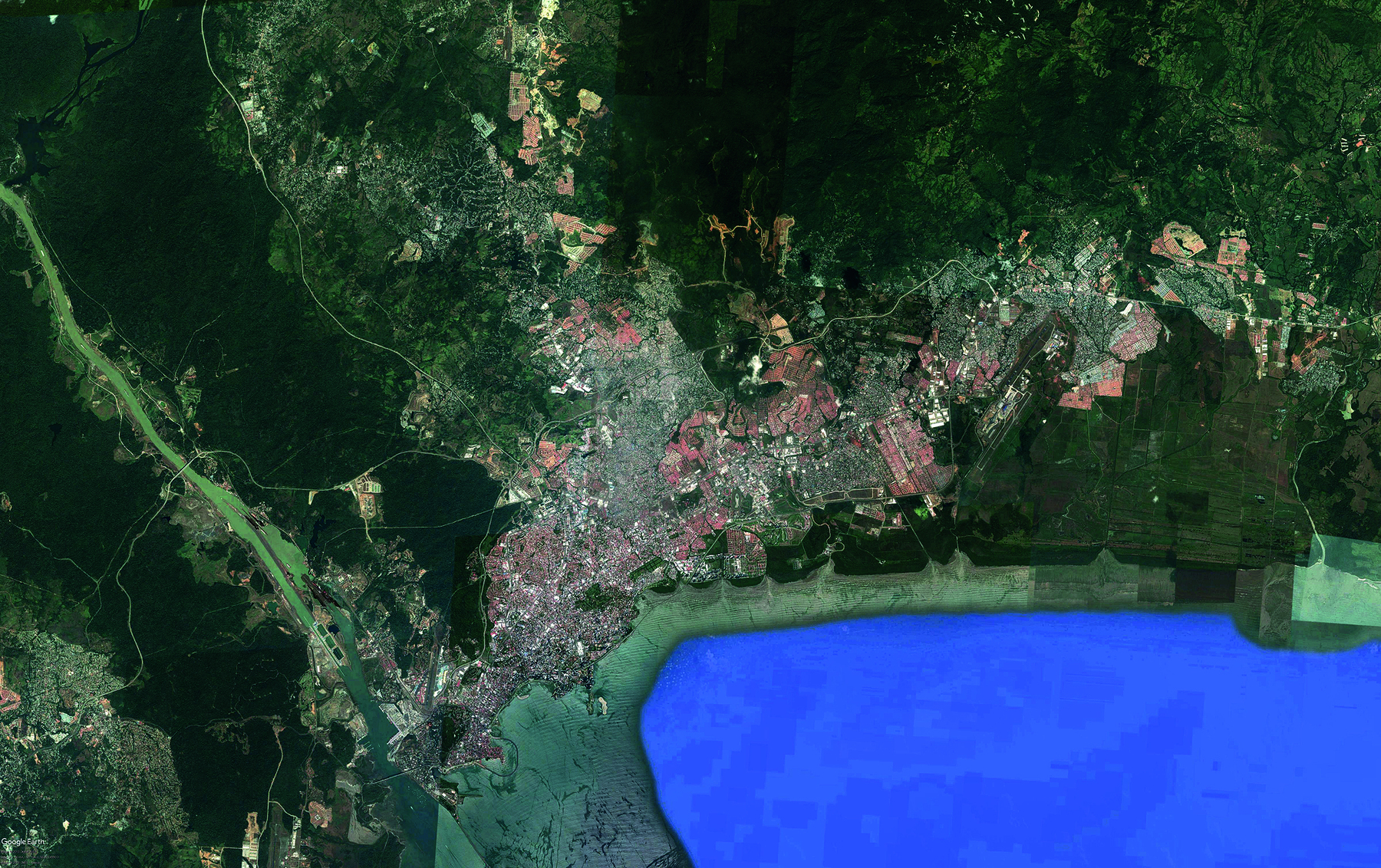 Imagem de satélite. A imagem representa uma porção territorial com grandes áreas verdes que misturam vegetação nativa e outras áreas de vegetação já alterada. Há trechos urbanizados pouco adensados nas bordas das áreas verdes e muito adensados na medida em que se aproxima de uma área com água, localizada do lado inferior direito da imagem. A área com construções estão aparecem em tons de cinza e marrom, sobretudo nas porções central e direita.