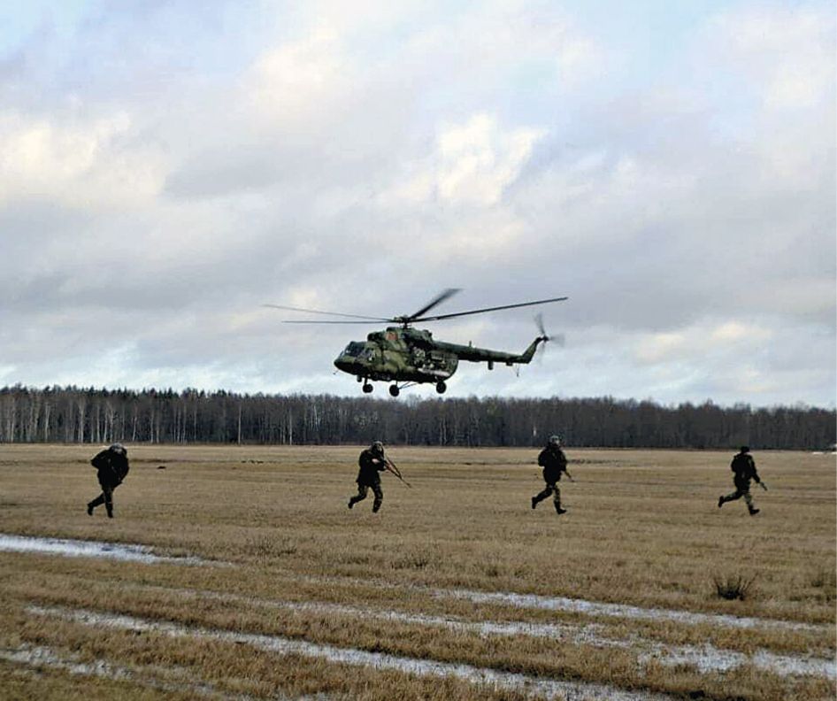 Fotografia.  Quatro soldados segurando armas correm em um campo aberto de vegetação marrom. Acima, há um helicóptero sobrevoando o campo. Ao fundo, diversas árvores.