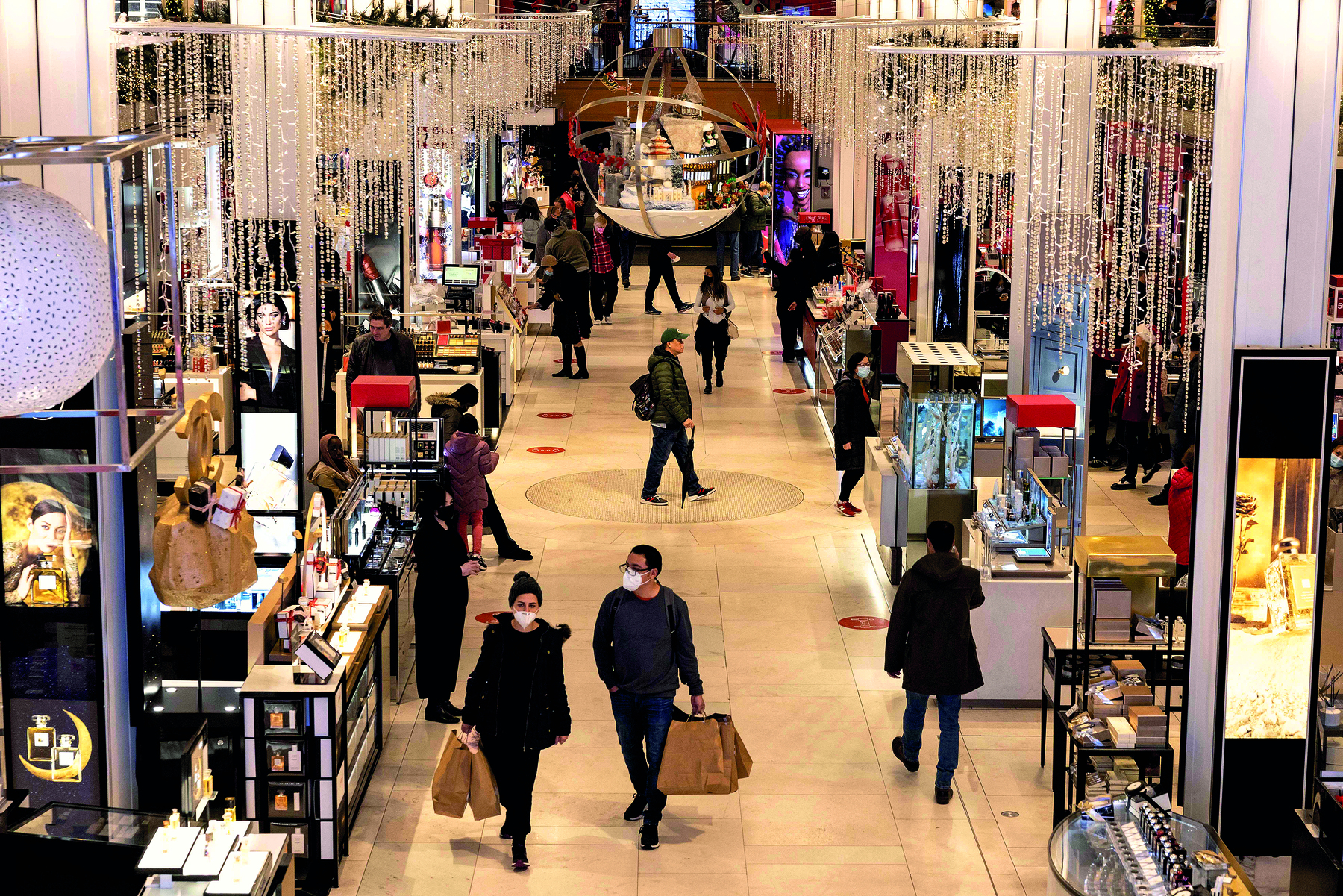 Fotografia. Vista do interior de uma loja; no centro há pessoas andando e carregando sacolas; nas laterais há diversos balcões iluminados, com produtos expostos e alguns atendentes.