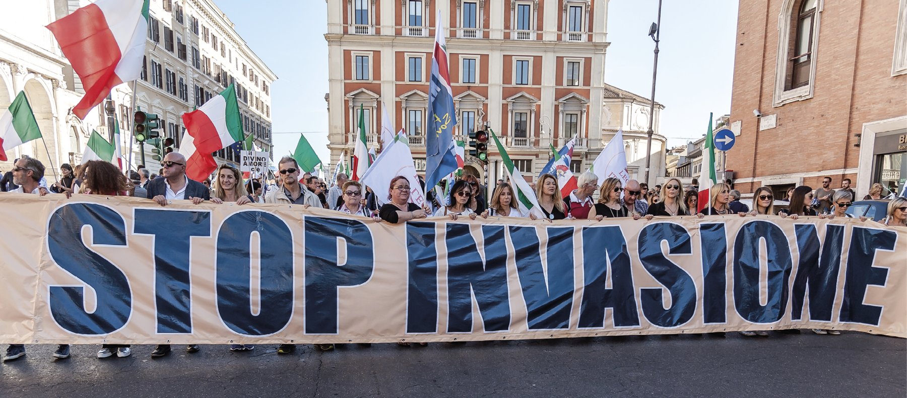 Fotografia. Diversas pessoas em pé segurando uma faixa com uma frase no idioma italiano. Ao fundo, há diversas bandeiras da Itália. Ao redor, prédios.