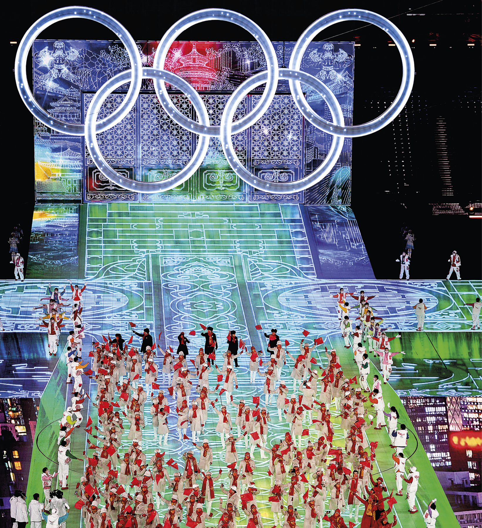 Fotografia. Vista de um palco com luzes nas cores azul e verde; em cima dele há diversas pessoas vestindo roupas brancas, vermelhas e pretas, segurando pequenas bandeiras vermelhas. No segundo plano, o símbolo das Olimpíadas, composto por cinco aros interligados.