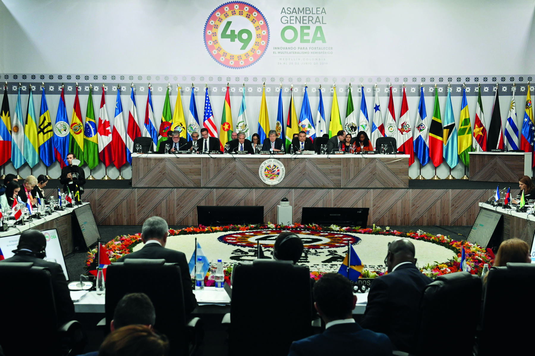 Fotografia. Vista panorâmica de uma reunião em ambiente fechado. No primeiro plano, algumas pessoas sentadas de costas para a foto, e tanto à direita quanto à esquerda, duas mesas ocupadas por outras pessoas. No segundo plano, diversas pessoas sentadas lado a lado, de frente para a foto e atrás de uma grande bancada marrom e branca. Ao fundo, há de diversas bandeiras hasteadas e, na parede, a sigla da Organização dos Estados Americanos.