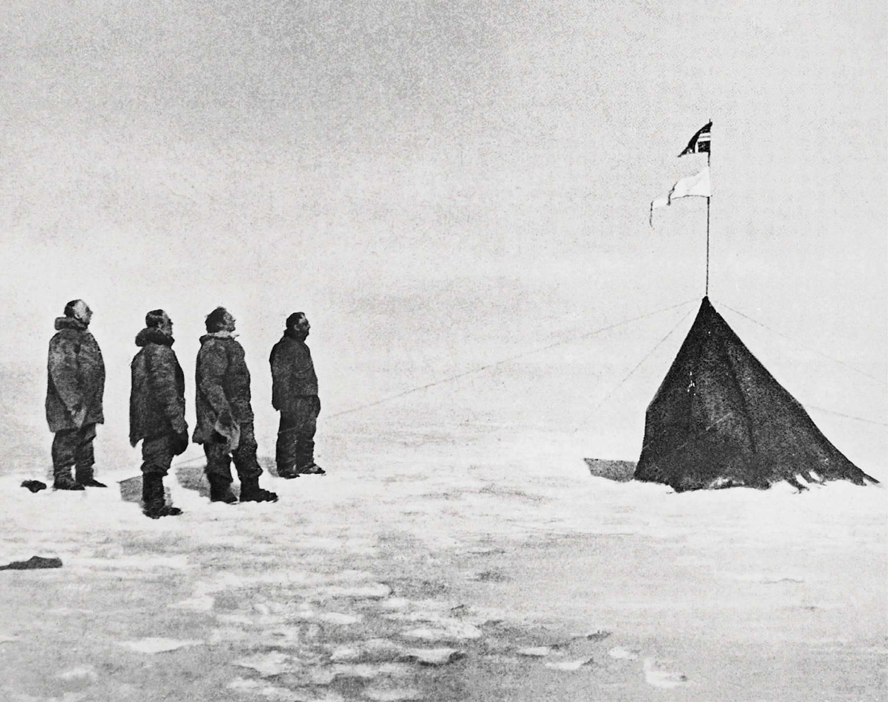 Fotografia.
Em preto e branco. Quatro homens agasalhados com casacos grossos e botas estão em pé sobre uma superfície coberta de neve, olhando para uma bandeira hasteada em mastro apoiado em uma tenda, à direita.