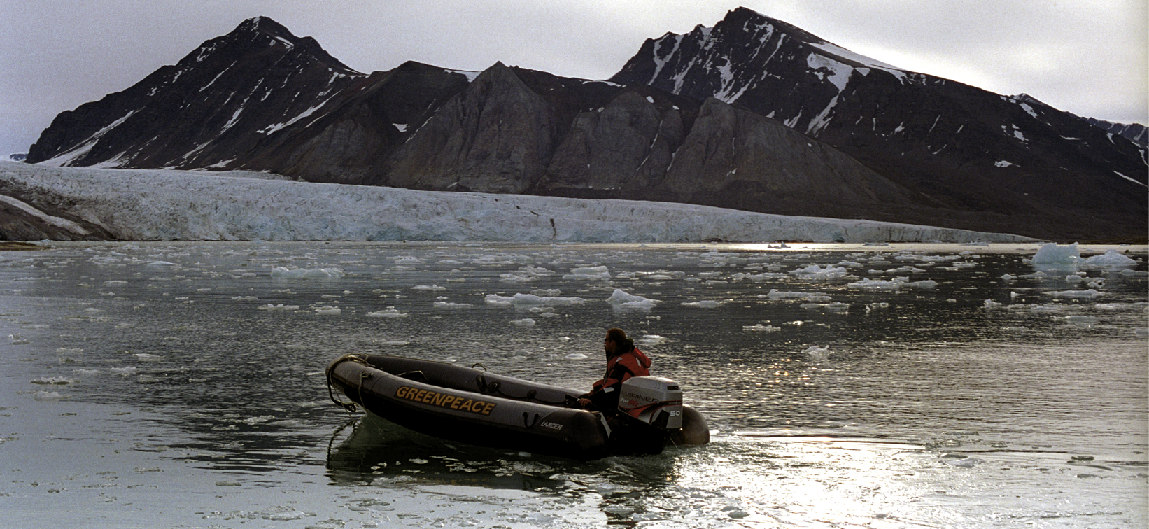 Fotografia. 
Uma pessoa dentro de um bote motorizado, em águas calmas, navegando  em direção a uma geleira baixa, ao fundo. Atrás da geleira, montes rochosos cobertos com pouca neve em algumas partes sob  céu nublado. Nas proximidades da geleira, pedaços de gelo flutuam sobre as águas.