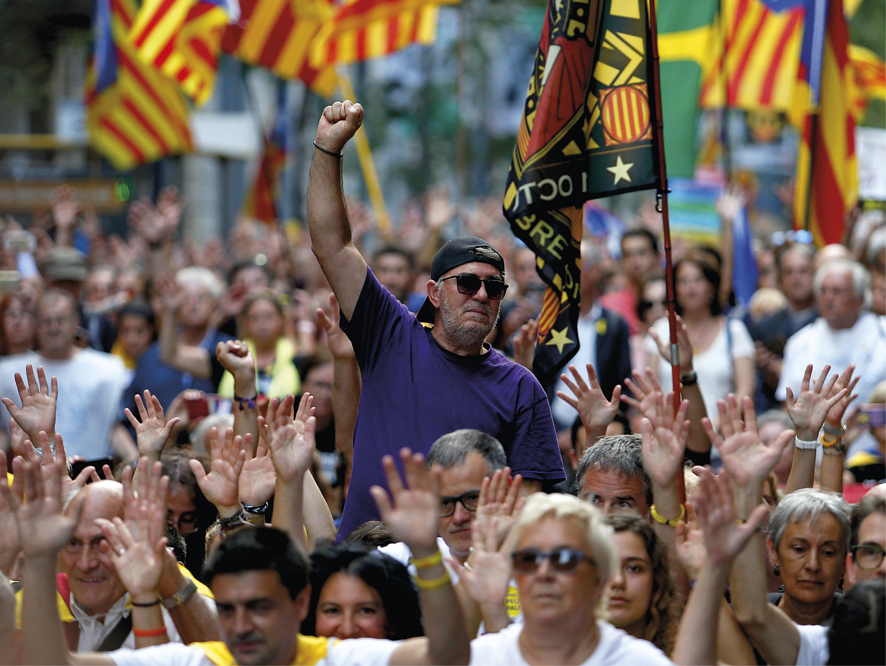 Fotografia. Multidão de pessoas andando com as mãos levantadas; no centro, destaque para um homem usando óculos escuros, boné, com a mão direita fechada e  levantada; atrás dele há diversas bandeiras com listras amarelas e vermelhas.
