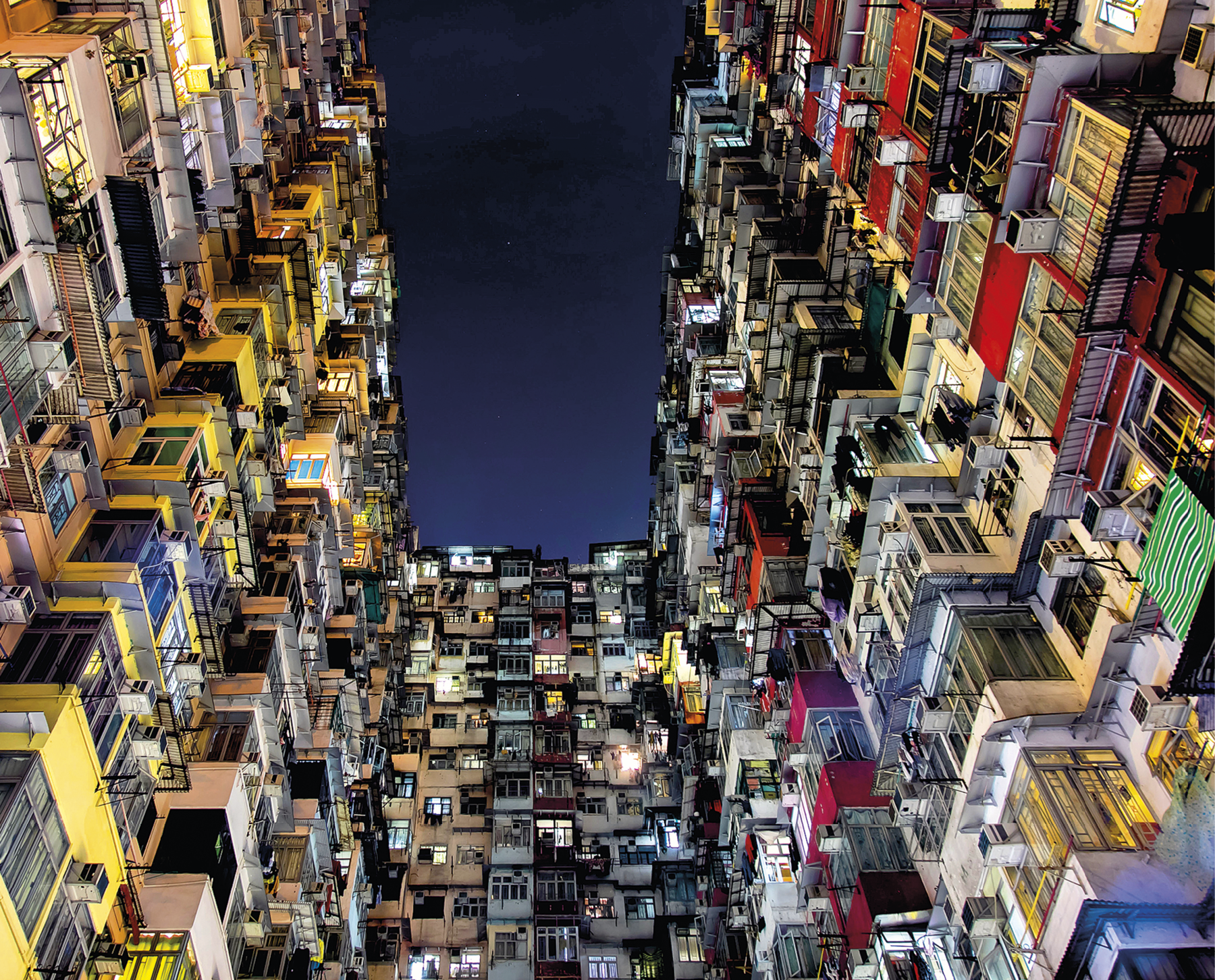 Fotografia. Vista noturna onde se observam vários prédios muito próximos uns dos outros. Há muitas janelas umas de frente para as outras.