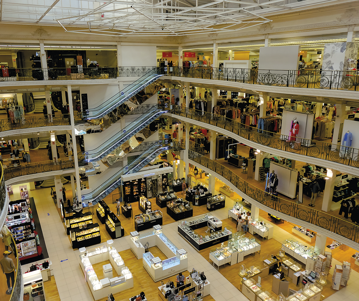 Fotografia. Interior de uma loja de departamentos com três andares, escadas rolantes, balcões com produtos no andar térreo, diversos manequins com roupas.