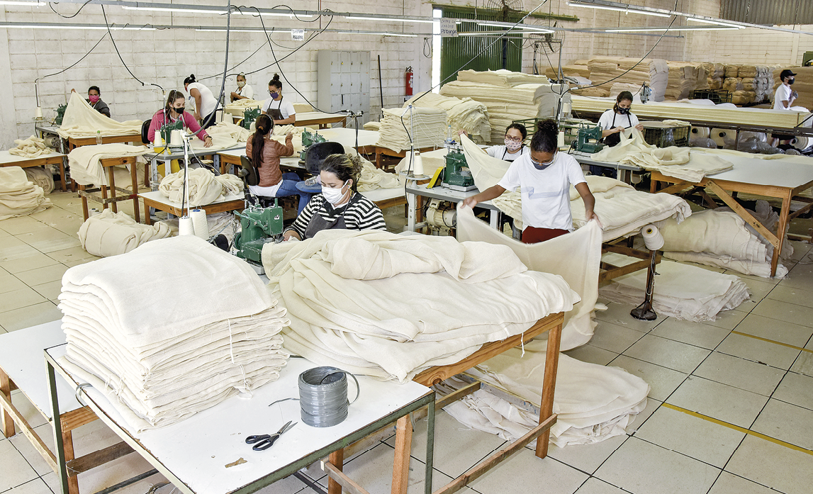 Fotografia. Diversas mulheres no interior de um galpão usando máscaras de proteção cobrindo o nariz e a boca. A maioria delas está em pé e ao lado de mesas com máquinas de costura e tecidos brancos empilhados.