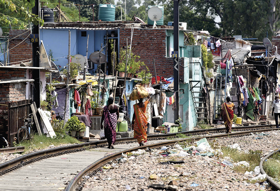 Fotografia. Duas mulheres usando vestidos longos andam sobre um trilho de trem. Uma delas carrega uma trouxa na cabeça. À esquerda do trilho há casas pequenas, precárias e muito lixo no chão.