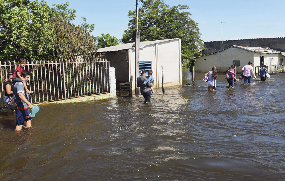 Fotografia. Diversas pessoas andam por uma rua inundada, com água até os joelhos; elas carregam crianças nas costas e no colo.