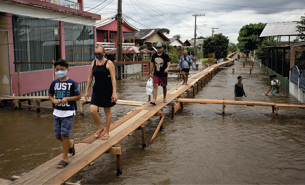 Fotografia. Pessoas usando máscaras de proteção cobrindo nariz e boca andam sobre uma pequena ponte de madeira construída sobre uma rua inundada. Ao redor há postes com fios e casas.