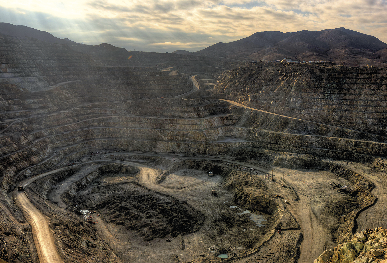 Fotografia. Vista de uma mina de cobre a céu aberto. A parte central da mina é a mais rebaixada, em tons de marrom e com estradas circulares de terra ao redor. Ao fundo, montanhas com vegetação.