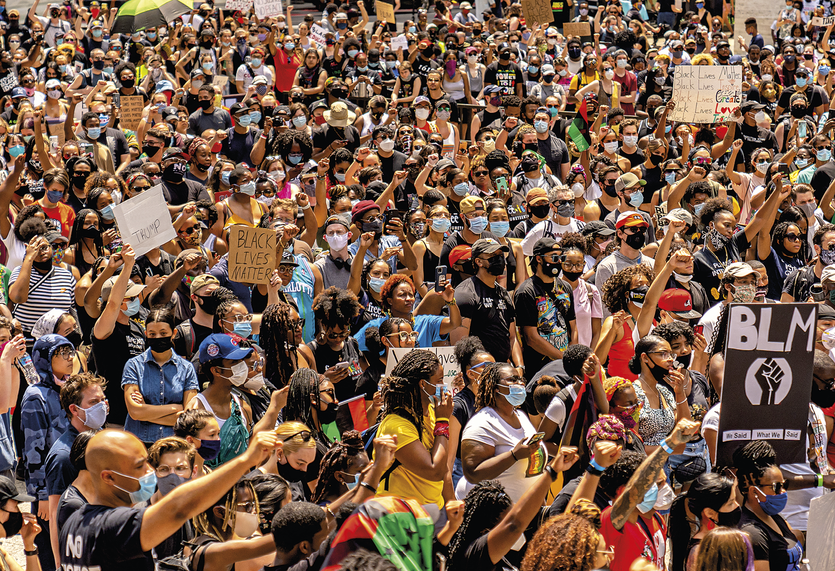 Fotografia. Uma multidão de pessoas reunidas em um dia de sol. Algumas delas usam máscara de proteção sobre nariz e boca; algumas pessoas estão com os braços erguidos e os pulsos fechados e outras seguram cartazes com os dizeres 'Black Lives Matter'.