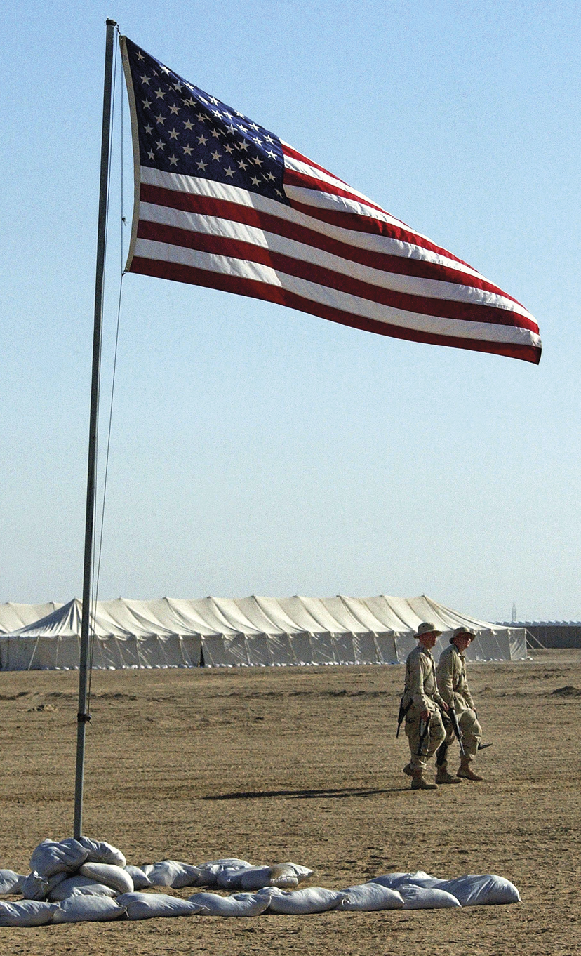 Fotografia. Área plana e aberta, de terra seca. No primeiro plano, bandeira dos Estados Unidos hasteada e tremulando e alguns sacos dispostos no chão. Atrás, dois soldados caminhando e, ao fundo, barracas brancas paralelamente enfileiradas. Céu azul.