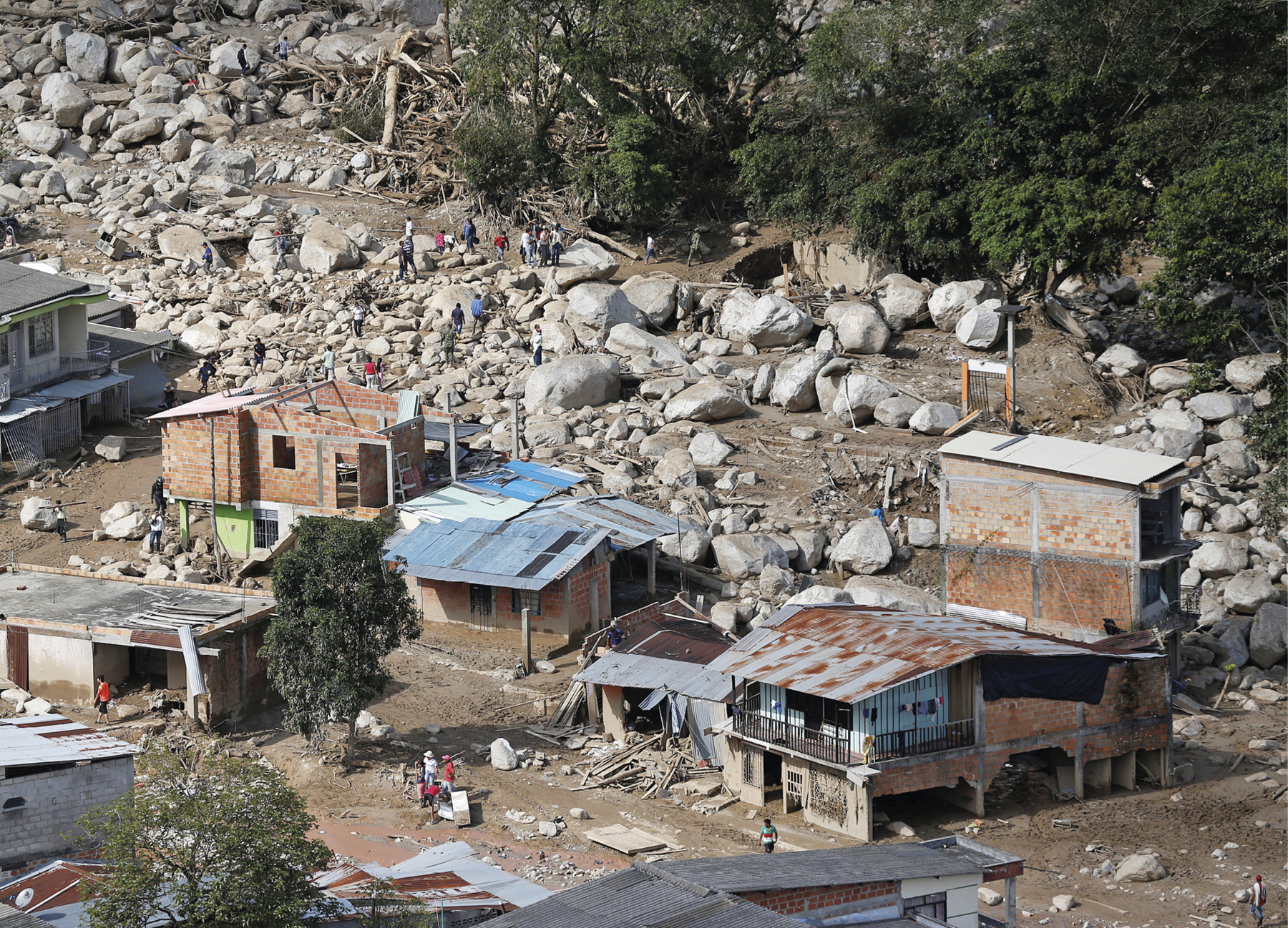 Fotografia. Ocupando a maior parte da imagem, área com chão de terra, algumas casas em condições precárias, sem acabamento e parcialmente destruídas, além de algumas pessoas caminhando em frente às construções. Ao fundo, diversas pedras brancas, aglomeradas e de diferentes tamanhos e pessoas circulando entre elas. Também ao fundo, do lado direito, árvores com parte dos galhos retorcidos.