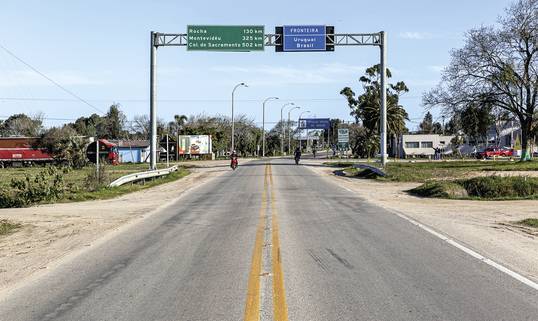 Fotografia. Vista do meio de uma rodovia de mão dupla na qual circulam poucas motocicletas. Ligada à uma estrutura metálica lateral, acima da estrada do lado direito, há uma placa azul em que está escrito: "Fronteira Brasil e Uruguai". Ligada à uma estrutura metálica lateral, acima da estrada do lado esquerdo, há uma placa verde em que está escrito: "Rocha 130 quilômetros, Montevidéu 325 quilômetros, Colônia do Sacramento 502 quilômetros." Ao redor da rodovia há várias árvores, acessos para caminhos de terra, algumas construções térreas e postes.