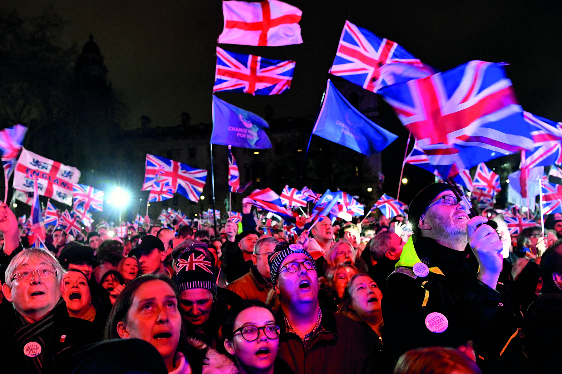 Fotografia. Uma multidão de pessoas reunidas em um espaço público aberto, durante a noite, vestindo agasalhos e gorros. Elas parecem estar cantando e algumas seguram a bandeira do Reino Unido e da Inglaterra.