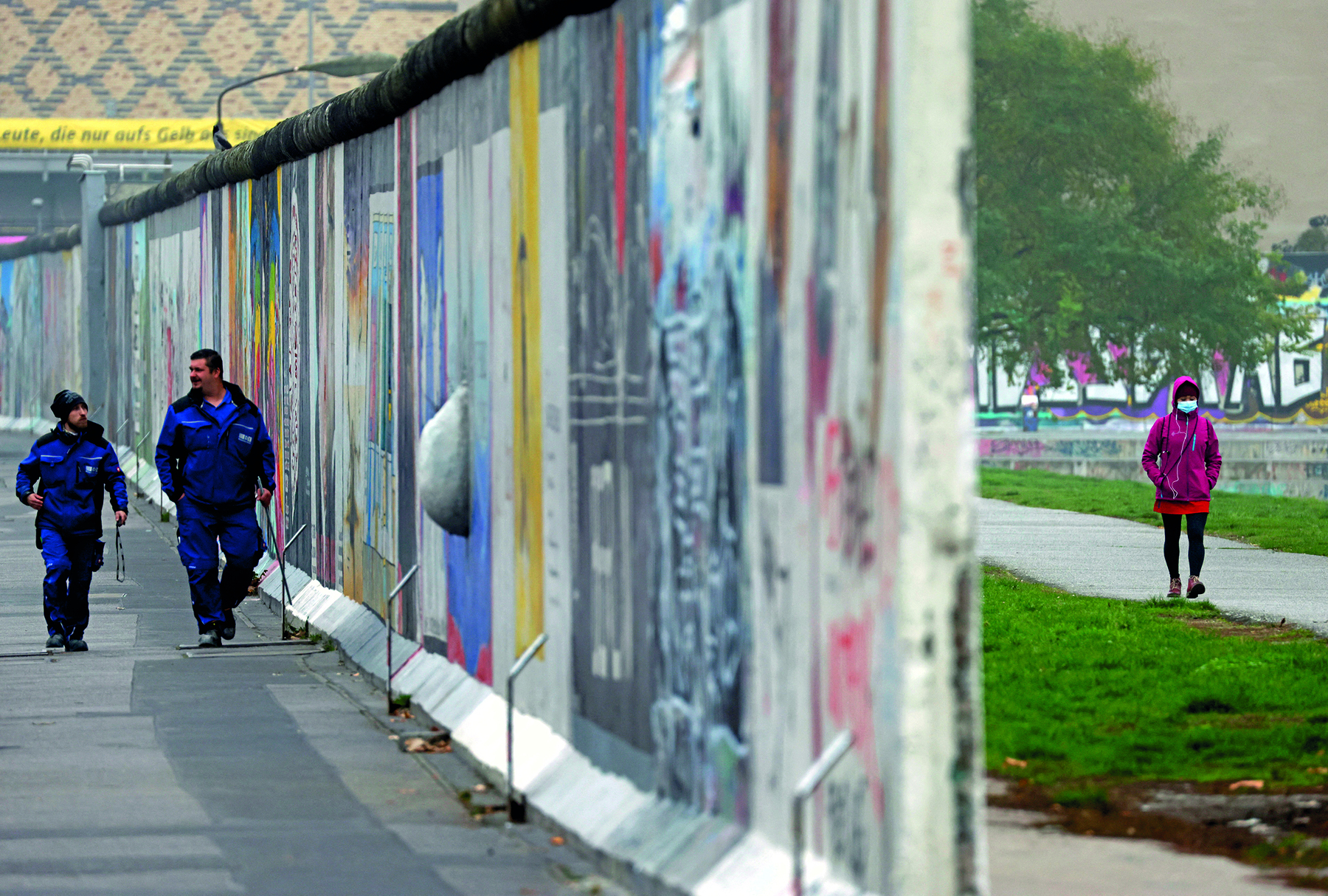 Fotografia. No centro da imagem, em destaque, o perfil de um muro alto e grafitado dividindo duas paisagens urbanas distintas. À esquerda do muro, duas pessoas vestidas com um uniforme azul caminham sobre uma calçada de concreto. À direita do muro, uma pessoa vestindo um agasalho rosa, máscara de proteção e gorro caminha com as mãos no bolso sobre uma calçada de concreto com gramado margeando à direita e à esquerda. 