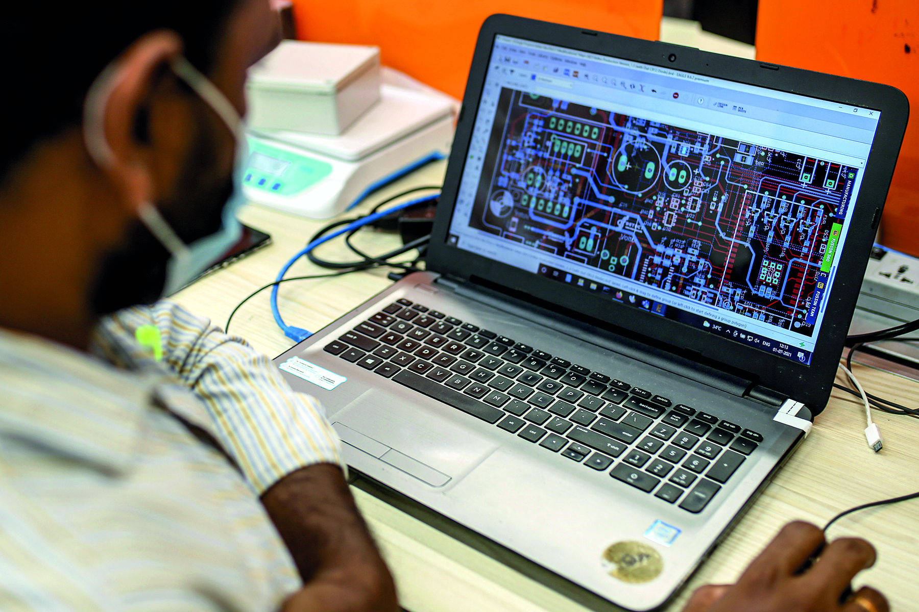 Fotografia. Destaque para um homem de barba e com máscara de proteção sentado em uma mesa em frente a um notebook. O homem manuseia o mouse e observa a tela do computador, que apresenta um projeto de tecnologia.