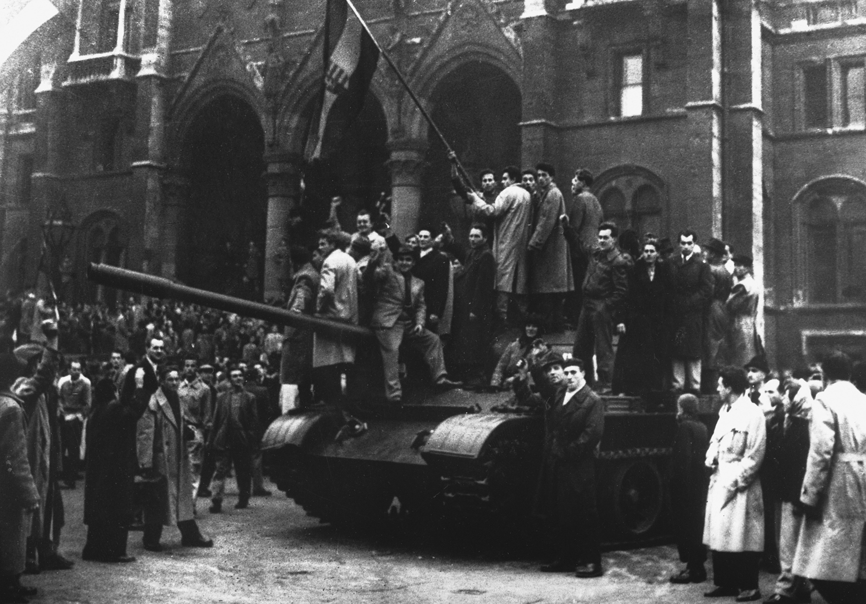Fotografia em preto e branco. No centro, diversas pessoas de casacos estão em pé sobre um tanque de guerra. Algumas delas seguram bandeiras. Ao redor do tanque, há uma multidão de pessoas em pé. Ao fundo, fachada de construções históricas.