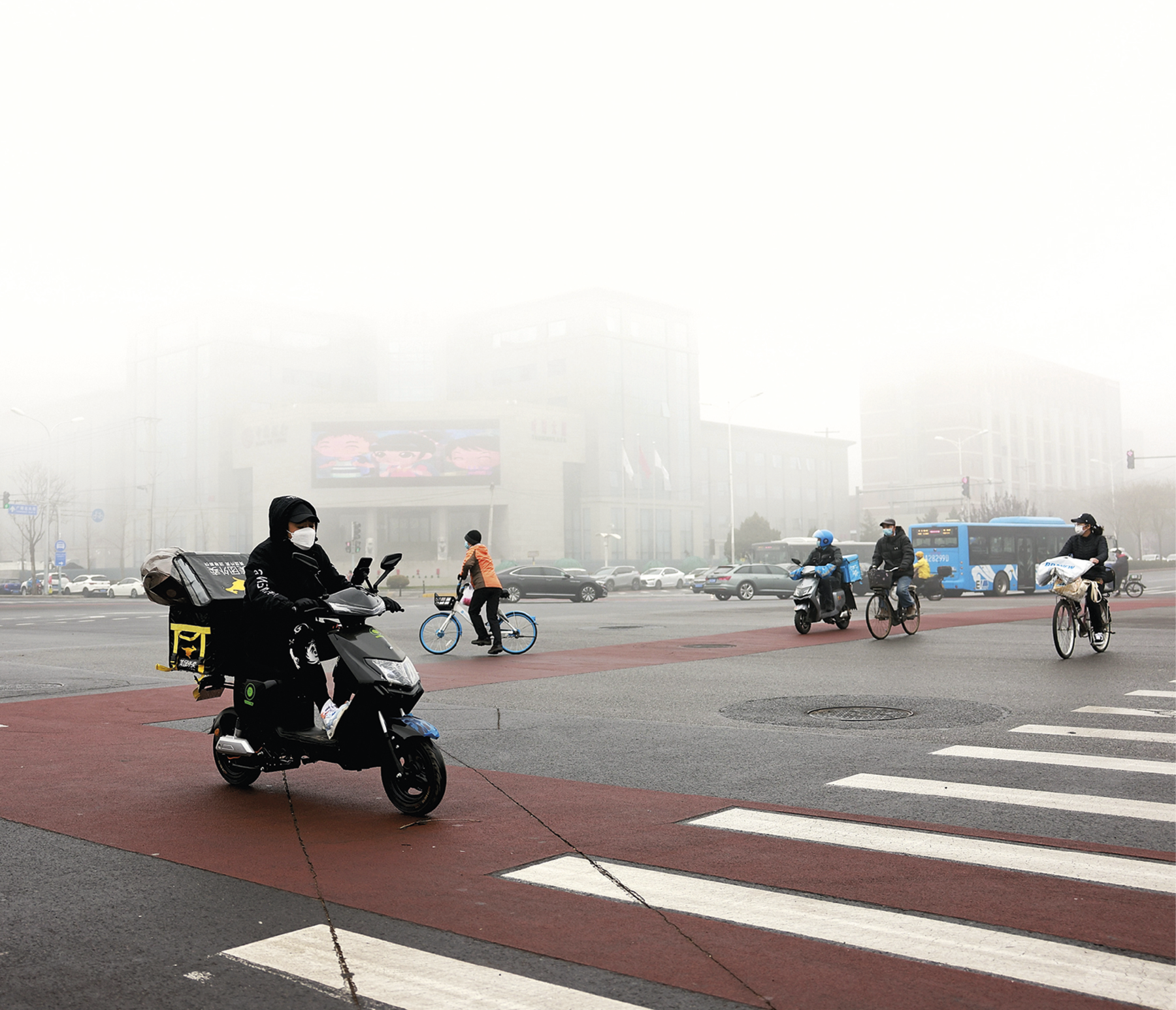 Fotografia. Vista para um cruzamento de avenidas. No primeiro plano, bicicletas e motocicletas cruzam a pista asfaltada e com a faixa da ciclovia sinalizada em vermelho. Os ciclistas e motociclistas usam máscara de proteção no rosto. No segundo plano, automóveis e ônibus parados no sinal vermelho e edifícios encobertos por uma densa camada de fumaça.